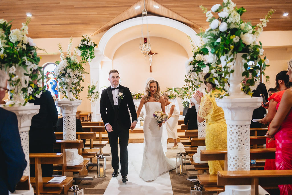 Bride and groom walking down a Leoni church aisle with white floral pedestals and lanterns