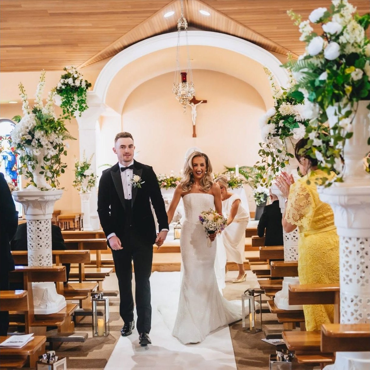 Bride and groom walking from the front row through a white floral church aisle