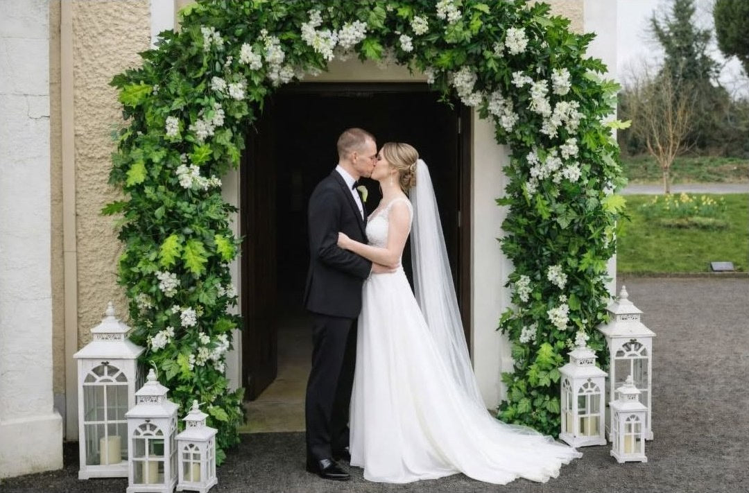 Bride and groom kissing beneath a green and white floral church entrance arch