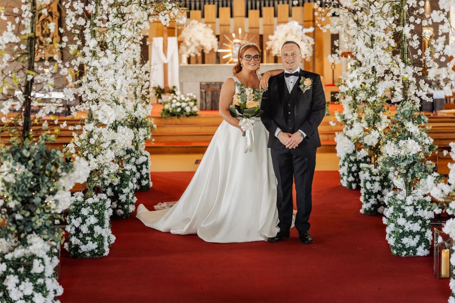 Bride and groom standing on a red church aisle surrounded by white wedding flowers
