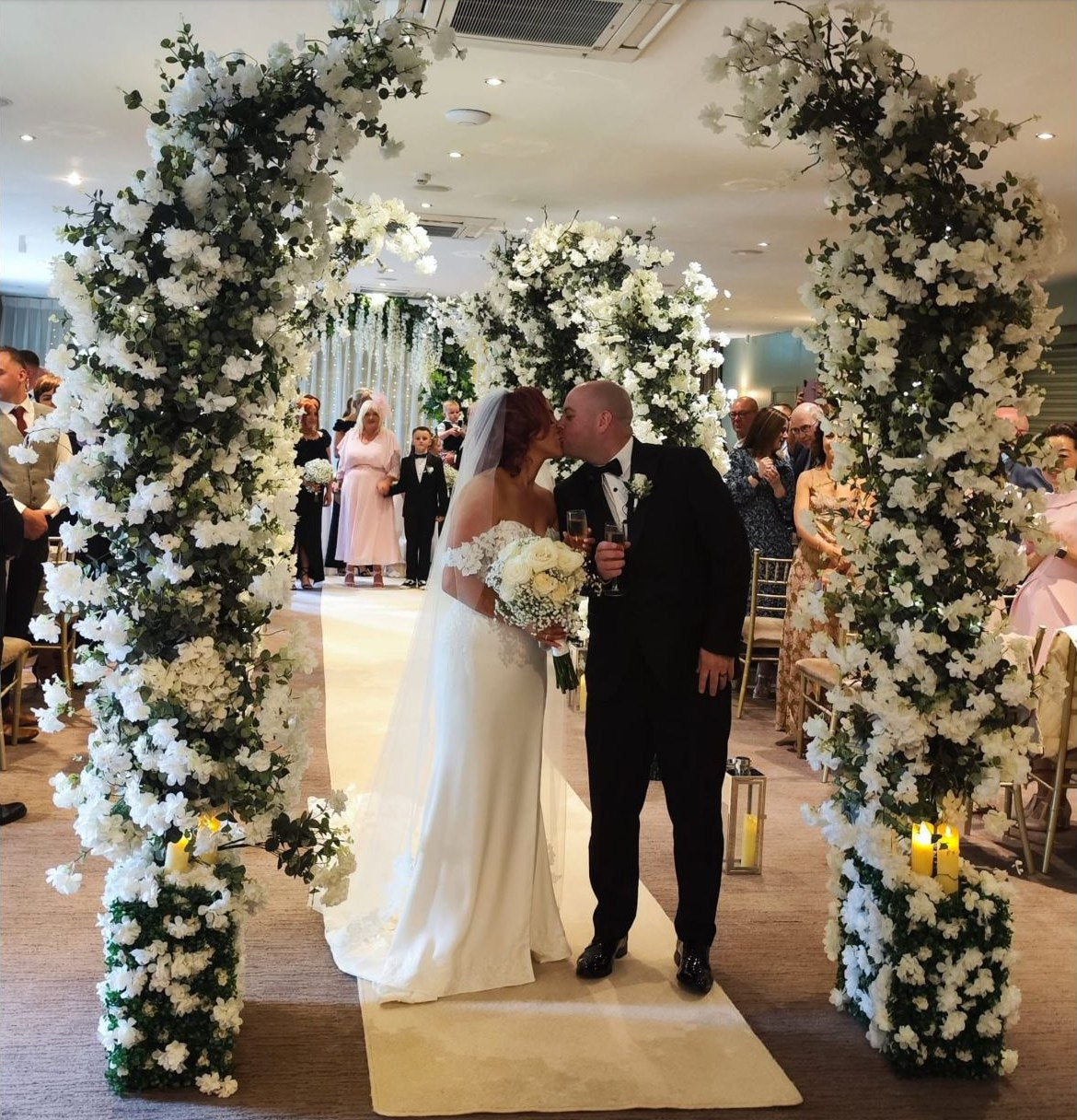 Bride and groom walking through a white blossom wedding aisle with guests nearby